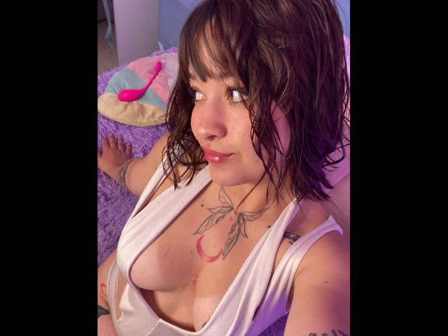 AnikaFleur with wet brown hair, white low-cut top, showing her boobs, tattoos on chest and arm, sitting on purple carpet, looking up with a slight smile.