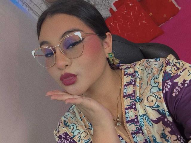 HelueJamil with glasses, pouty red lips, and colorful patterned top, blowing kiss. She's got green earrings and a gold necklace, looking cute and stylish.