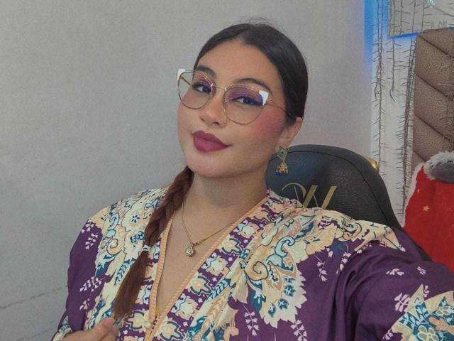 HelueJamil with dark hair in a braid, wearing glasses, purple and gold patterned top, red lipstick, and gold earrings, smiling slightly, holding her right breast.