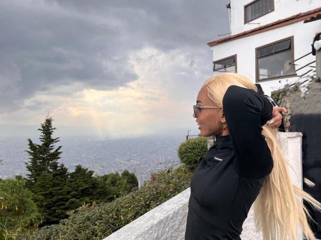 AylaCallowey with long blonde hair, black long-sleeve top, and glasses, standing with arms behind head, looking at distant city, next to white building with trees in foreground.