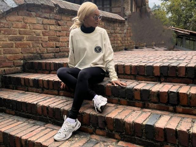 AylaCallowey with blonde hair, glasses, white sweater, black turtleneck, black pants, white sneakers, sitting on brick steps, looking to the side.