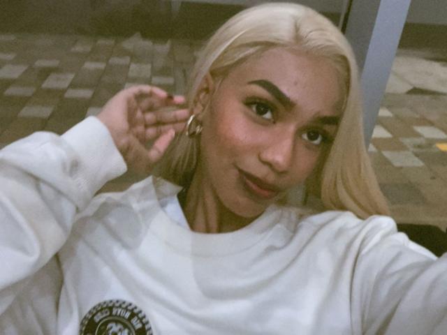 AylaCallowey with blonde hair, wearing a white sweatshirt, gold hoop earrings, and a slight smile, holding her hair up. Her tits are small, and she has dark eyebrows.