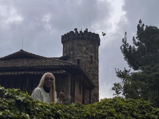 AylaCallowey with long white hair, wearing a white top, stands in front of a stone castle with a tower, surrounded by greenery and a cloudy sky.