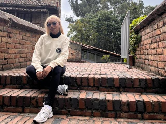 AylaCallowey sits on brick steps wearing a white sweater, black pants, and white sneakers, with long blonde hair and sunglasses, in a rustic outdoor setting.