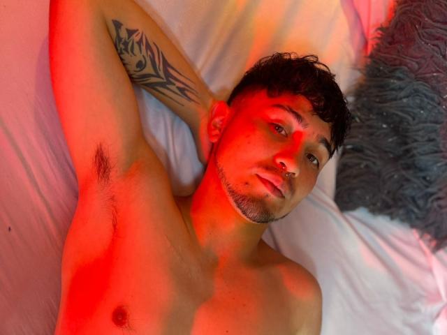 ScarlettAndAlan, shirtless, with red lighting, dark curly hair, tattoo on arm, nose ring, armpit hair, laying on bed, looking at camera, white pillow, gray fuzzy pillow.