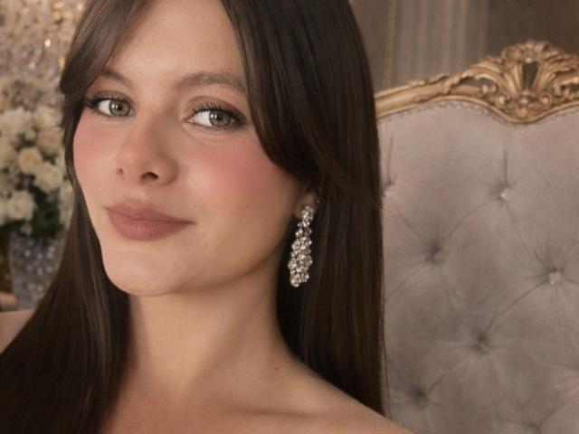 VictoriaWaynes has long brown hair, green eyes, and a subtle smile. She's wearing sparkly earrings and has a slight blush on her cheeks. Her boobs are not visible.