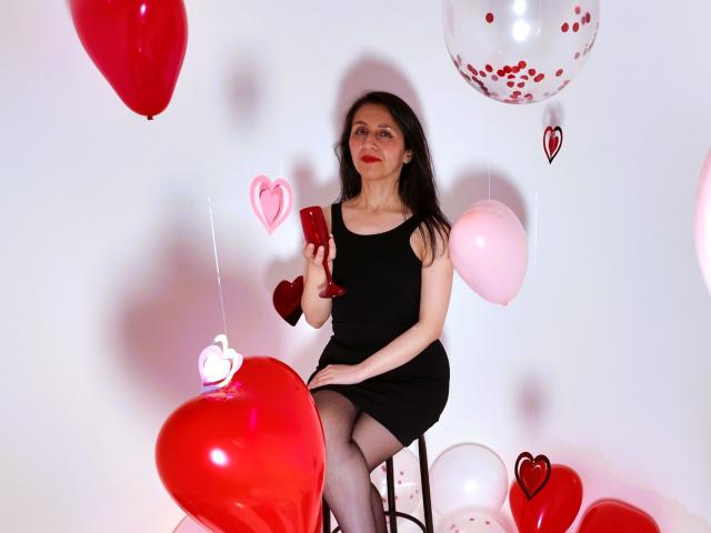 Cyntiia with long dark hair, red lipstick, black dress, and black stockings, holding a red glass, sitting on a stool surrounded by red and pink heart balloons.