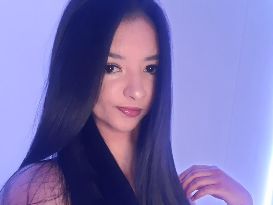 MaraGreey with long black hair, slightly smiling, wearing a black top, right hand touching hair, blue light background, shiny pink lipstick, light skin, subtle makeup.