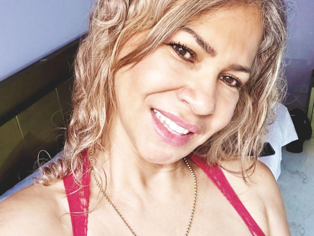 BrigitteWalsh with curly blonde hair, pink lipstick, and red tank top, smiling at the camera, showing off her white teeth and gold necklace. Her tits are partially visible.