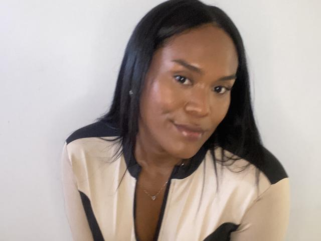 AshleyRob has dark skin, long black hair, and is wearing a beige and black shirt with a small necklace. She's smiling slightly with a relaxed expression.