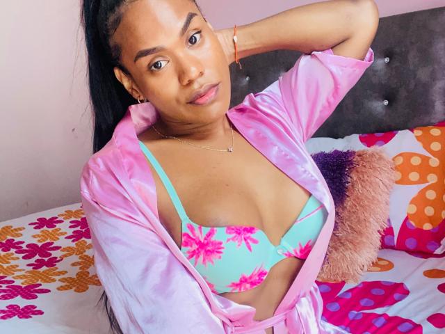 AshleyRob with brown skin, black hair in a ponytail, wearing a pink silk robe and a turquoise floral bra, showing her tits, sitting on a colorful bed.