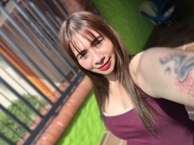 MelannyTaylorr with red lipstick, brown hair, and a tattoo on her arm, smiling in a purple tank top, showing her tits in a selfie. Bright green wall in background.
