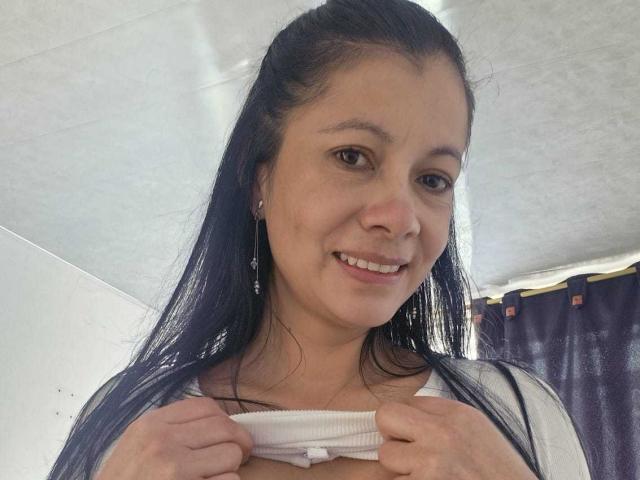 MelissaBaud with long black hair, smiling, pulling up a white ribbed top to reveal her small, perky tits. She has light skin and brown eyes.