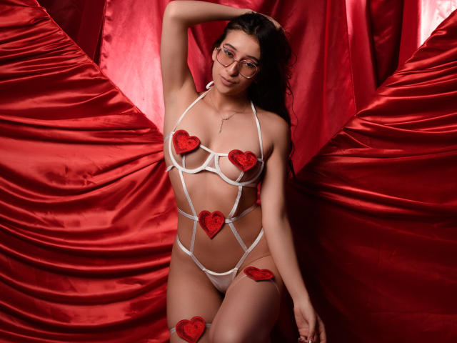 Paulla69x in white strappy lingerie with red heart pasties covering her tits and pussy, wearing glasses, against red satin. Her right arm is raised, showing off her tits.
