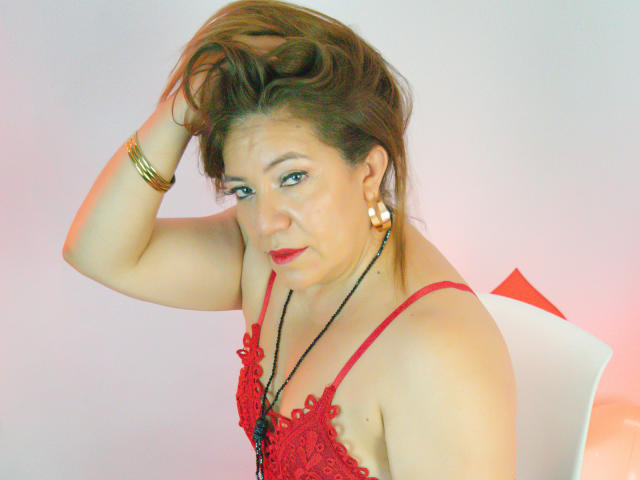 JadeSexiest, a middle-aged woman with light brown hair, sits with one hand in her hair. She wears a red lace spaghetti-strap top, gold hoop earrings, and a black necklace. Her red lipstick contrasts with her fair skin. She has a slightly annoyed expression. The image has a soft, pastel-colored background with a white chair and red triangle. Her bracelet is gold. The photo has a slightly grainy texture.
