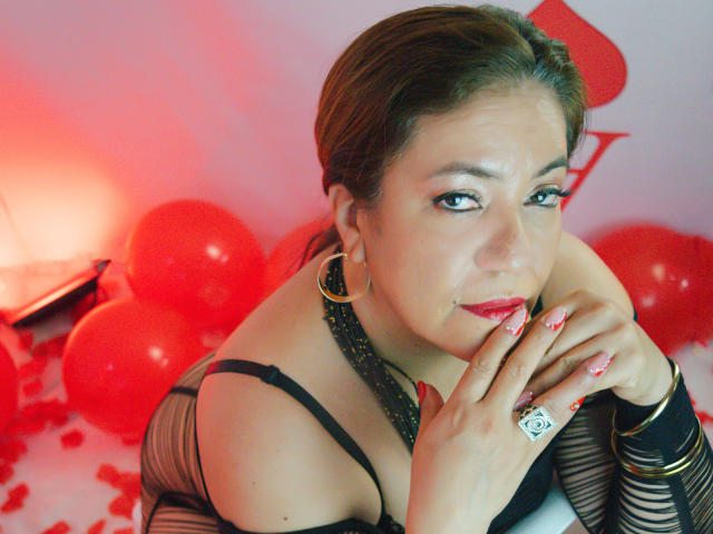 JadeSexiest is a medium-skinned woman with brown hair, wearing black fishnet lingerie and large gold hoop earrings. She has red lipstick and eye makeup, with red-painted nails. She's leaning forward, looking at the camera with a slight smile. Her right hand is near her face, showing a ring with a white square symbol. Red balloons and petals are scattered around her. The image has a red and white color scheme.