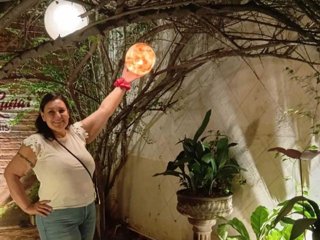 LadySelene with a fat orange orb in her right hand, wearing a white shirt and blue pants, standing under a vine-covered arch with a plant to her right.