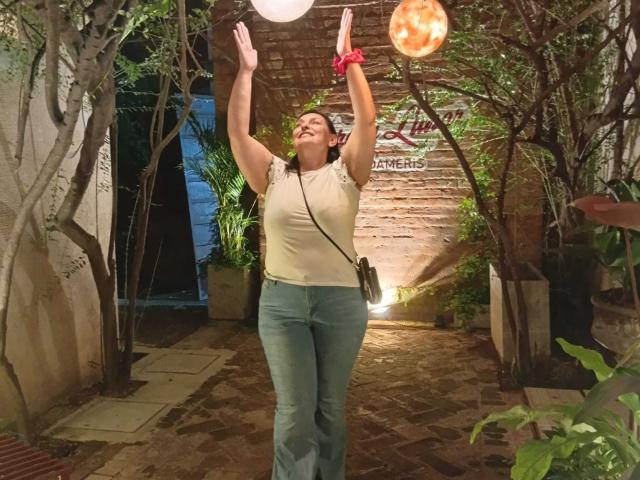LadySelene with arms raised, white shirt, blue pants, red scrunchie, and black crossbody bag, smiling at a glowing orange balloon in a nighttime garden setting.