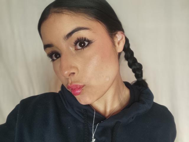 EmilyVv has dark brown eyes, long black braid, pink lipstick, and wears a black hoodie. Her pouty lips and subtle blush are visible. Her skin is light brown.
