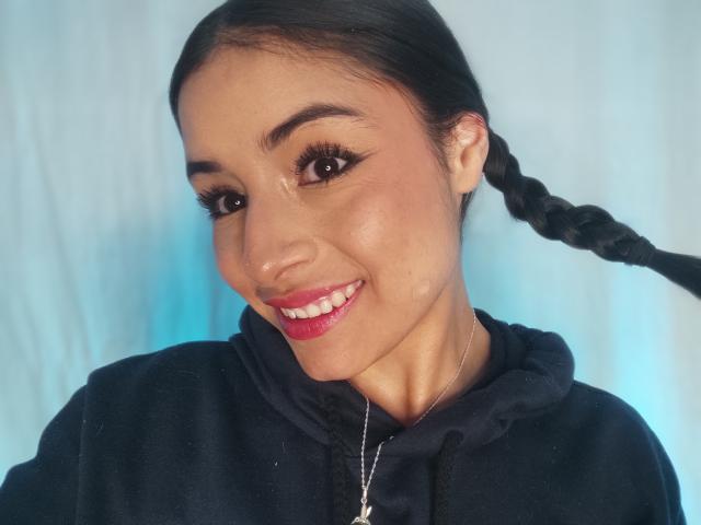 EmilyVv with long black braid, wearing a black hoodie, smiling with red lipstick, brown eyes, and subtle makeup, has a small silver necklace against a blue backdrop.