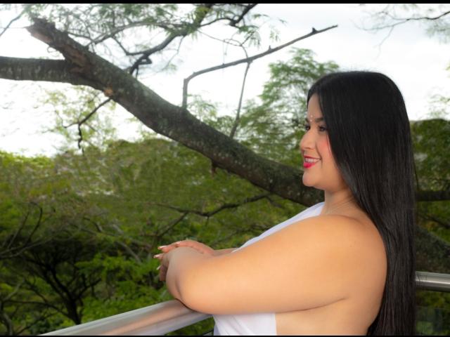A photograph of MiahKumarI with long black hair, smiling, facing right. She has a curvy, full-figured body with light brown skin. She wears a white, sleeveless top. Her arms rest on a metal railing. A large, dark tree branch extends diagonally behind her, with green foliage in the background. Bright, natural light highlights her face and hair. The composition focuses on MiahKumarI's profile and the tree branch.