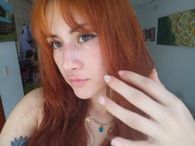 MalenaJones with red hair, nose ring, lip piercing, and green necklace, touching her face, showing tattoos and pale skin, blue eyes, and slightly parted pink lips.