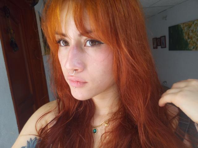 MalenaJones with red hair, nose ring, lip piercing, green necklace, and tattoo on shoulder, looking at camera, holding hair, pale skin, brown eyes, slight pout, indoors, sunlight.