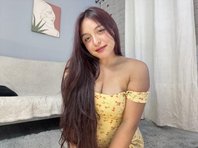 MarianaVargasX with long brown hair, yellow floral off-shoulder dress, small tits, cute smile, sitting, white curtain, white sofa, light blue wall, flower painting in background.