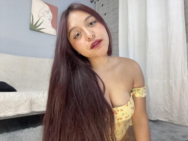 MarianaVargasX with long brown hair, wearing a yellow floral off-shoulder top, showing her boobs, nose ring, and red lipstick, sitting in a white-walled room.