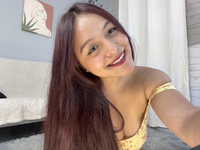 MarianaVargasX with long brown hair, smiling, wearing a yellow off-shoulder top, showing her small tits, nose ring, and red lipstick, taking a selfie.