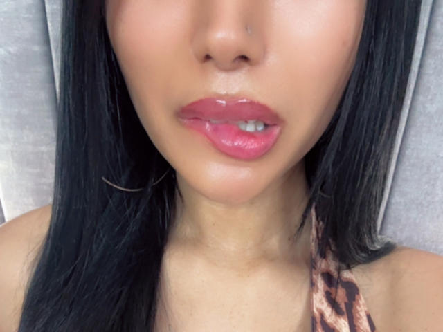 Close-up of VanezzaTss's face. She has smooth, tan skin and full, glossy pink lips. Her black, straight hair falls on both sides of her face. She wears a small, visible beauty mark near her right upper lip. Her visible teeth are slightly parted. She has a small silver nose ring on her right nostril. She wears a leopard-print halter top, and the background is a soft, grey texture. The focus is on her lips and facial features.