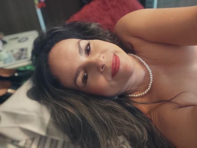 Danny69 está acostada en una cama, con cabello largo y oscuro, lápiz labial rojo, un collar de perlas, sonriendo a la cámara, sin blusa, mostrando sus senos, ojos marrones, piel clara..