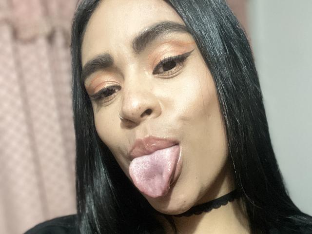 StellaBeckker has long black hair, a nose ring, and is sticking out her pink tongue with her lips slightly parted. She's wearing orange eyeshadow and a black choker.
