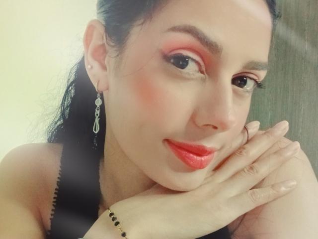 AnaisHilton with red makeup, red lipstick, black hair in a ponytail, earrings, touching her chin, smiling, wearing a black top, and a bracelet on her right wrist.