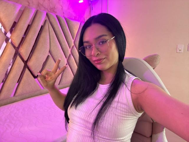 IsaDoux with long black hair, glasses, and white ribbed tank top, making a peace sign, pink and purple lighting, sitting in a chair, showing her tits.