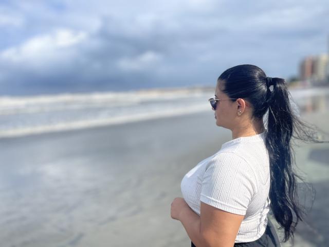 MiahKumarI with long black hair in a ponytail, wearing sunglasses and a white ribbed shirt, showing her big tits, standing on a beach with waves in the background.