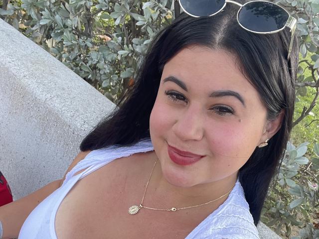 Close-up of MiahKumarI with medium brown skin and black hair. She's wearing a white top with a low neckline, showing a bit of cleavage. She has dark eyebrows, long eyelashes, and red lipstick. Her earrings are small and gold. A gold necklace with a small flower pendant rests on her chest. Sunglasses sit on her head. She's smiling slightly. The background has green and gray foliage. The image has a natural, outdoor light.