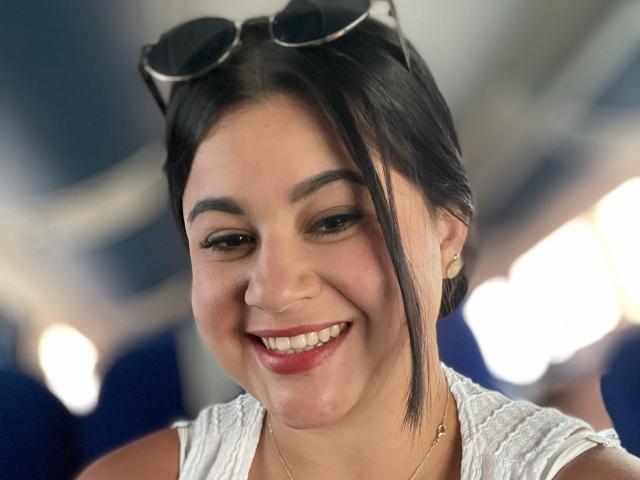 Close-up portrait of MiahKumarI with light brown skin, dark straight hair in a side part, and a small nose. She's smiling with red lipstick, white teeth, and small earrings. Eyeglasses rest on her head. She wears a white lace shirt. Blurred blue and white background. Image focuses on her face. Textures: smooth skin, lace shirt. Colors: light brown skin, dark hair, white lace, red lipstick. Slight shadow on her cheek. Natural light. MiahKumarI looks relaxed and happy.