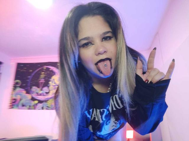 AshGraven with long blonde and brown hair, black lipstick, tongue piercing, and black eye makeup, sticking tongue out, making a peace sign, wearing a black Vampire rock shirt.