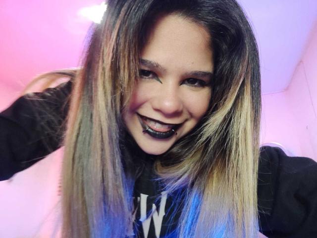 AshGraven with long, straight blonde and brown hair, black lipstick, and a big grin, wearing a black hoodie, in a pink-lit room, looking directly at the camera.