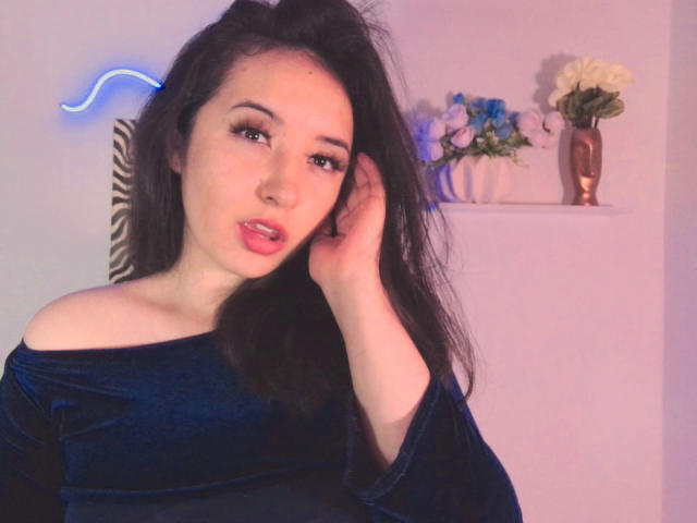 SusanMonroy with long black hair, wearing a blue velvet off-shoulder top, touching her hair, pink lipstick, neon blue light in background, white flowers on shelf.