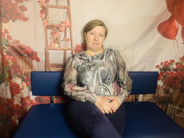 RadaChudna sits on a blue bench, wearing a patterned blouse with "boobs" and dark pants, hands on lap, looking straight at camera, with short brown hair.