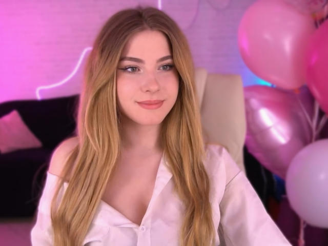 SabrinaMuse with long blonde hair, wearing a white shirt, smiling softly, pink and purple lighting, pink and white balloons, her tits partially visible, sitting in a chair.