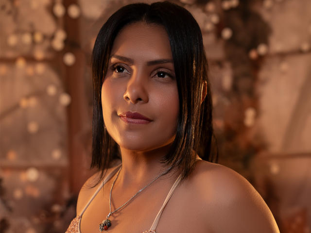 BarbaraSweetty with shoulder-length black hair, medium skin, and subtle makeup, wearing a necklace, smiling softly, with warm, soft lighting highlighting her face and upper body.