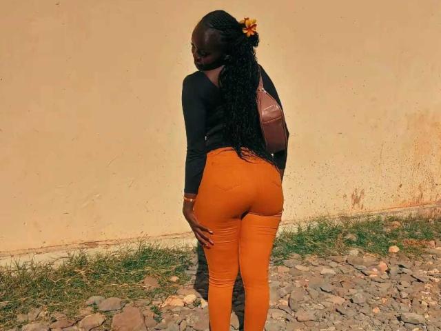 Jadah69 with dark skin, long braided hair, black top, orange tight pants, brown backpack, standing on rocky ground, looking back, showing her ass.