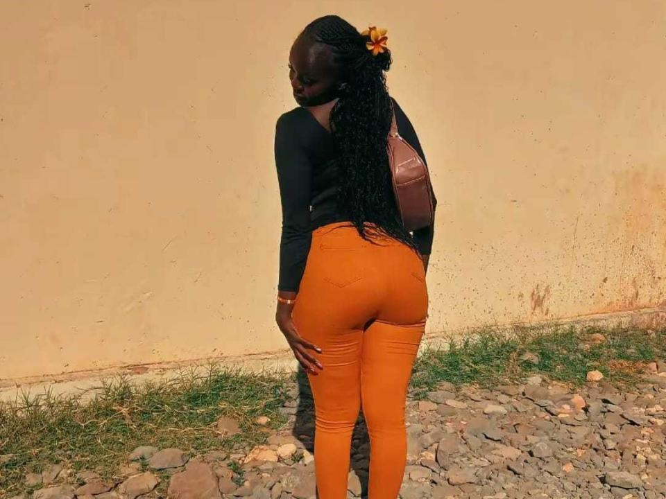 Jadah69 with dark skin, long braided hair, black top, orange tight pants, brown backpack, standing on rocky ground, looking back, showing her ass.