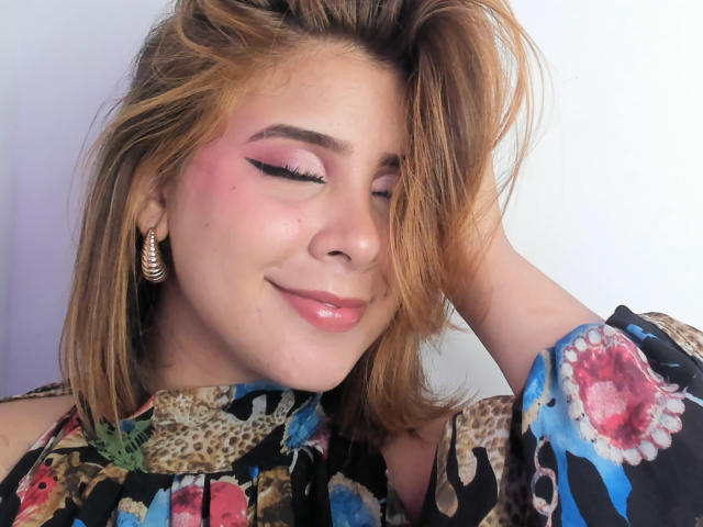 ZaraVidal with pink eyeshadow, glossy pink lips, and a slight smile, wearing a colorful floral top, hand in messy brown hair, gold earrings, and a sexy, cute face.