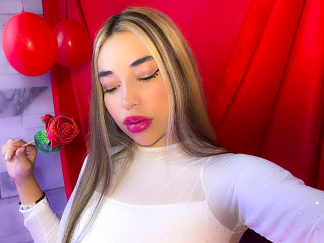 MissVallen with long blonde hair, pink lipstick, and white sheer top, holding a red rose lollipop, has pink eyeshadow, and a cute, sexy look, standing in a red background.