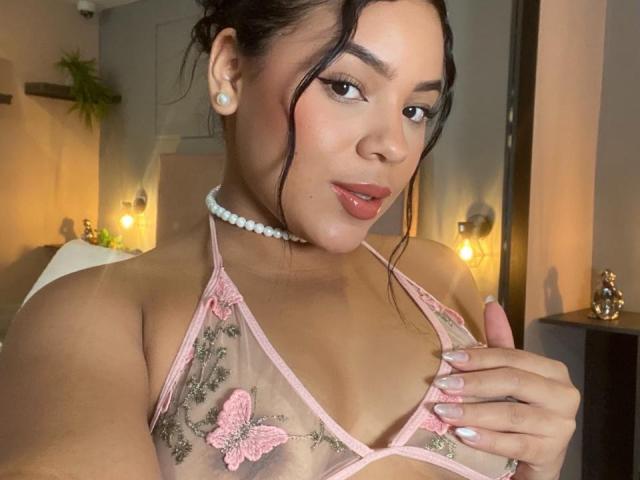 AuroraaRose with brown eyes, pink lips, and dark hair in a half-up style, wearing a sheer pink lace bra, pearl necklace, and earrings, holding her small tits.