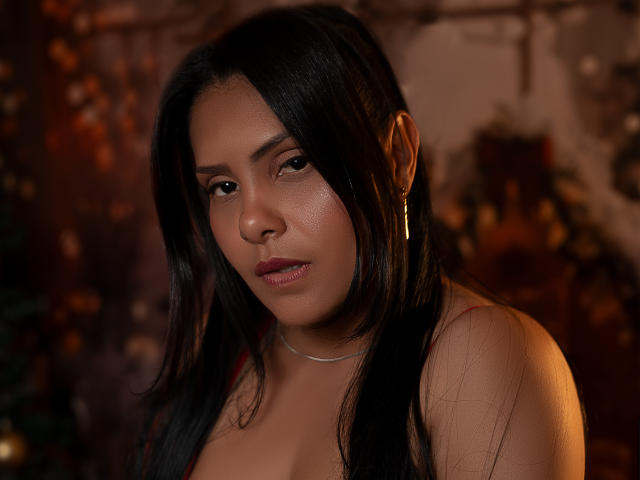 BarbaraSweetty has dark, straight hair, brown eyes, and a slightly flushed face with glossy pink lips. She wears a gold earring and a red top, showing her tits.